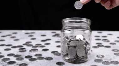 Hand Throws One Five-Kopeck Ukrainian Coin into a Glass Jar Filled with Coins. A Pile of scattered coins is on table. Stock. Piggy bank. Black background. Financial crisis, budget savings. Poverty.