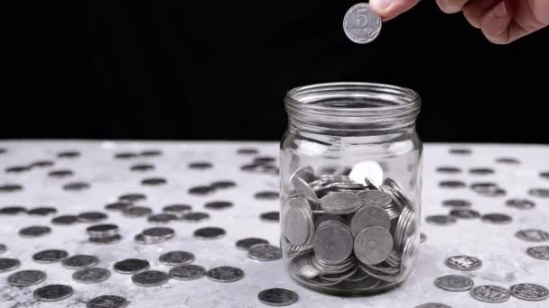 手把一个五科普克乌克兰硬币扔进一个装满硬币的玻璃瓶里 桌子上有一堆散落的硬币 小猪银行 黑色背景 金融危机 预算节约 — 图库视频影像