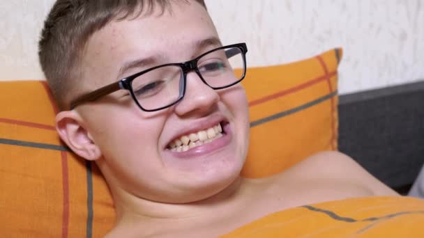 一个带着眼镜躺在床上的青少年 做鬼脸 露出牙齿 鬼鬼祟祟的 累了的男孩休息 改变面部表情 和乐趣 用孩子的牙齿咧嘴笑 — 图库视频影像
