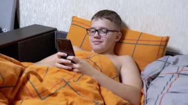 Gözlüklü Çocuk, rahat bir yatakta uzanan akıllı telefondan ilginç bir video izliyor. Gülümseyen çocuk, turuncu bir battaniyeyle kaplı bir yastığa uzanarak rahatlıyor. Yatma vakti. İnternet bağımlılığı. Çevrim içi sohbet.