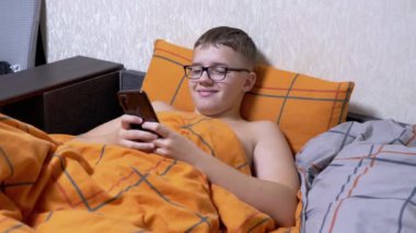 Gözlüklü Genç, Smartphone 'da mesaj yazıyor, rahat bir yatakta yatıyor. Gülümseyen çocuk, turuncu bir battaniyeyle kaplı bir yastığa uzanarak rahatlıyor. Yatma vakti. İnternet bağımlılığı. İnternette. Video oyunu.