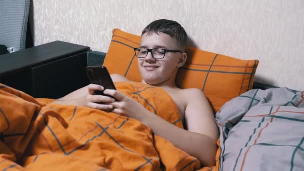 眼镜中的青少年在舒适的床上躺在智能手机上写短信 笑着的男孩放松了下来 躺在枕头上 上面铺着一条橙色的毯子 该睡觉了上网成瘾 电子游戏 — 图库视频影像