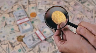 Bitcoin Sikkenin Para Arkaplanındaki Büyüteç ile Macro Görünümü. Elinde bir bitcoin tutan bir kadın, ve onu arıyor ve inceliyor, bir loupe kullanarak. Masaya 100 dolarlık banknotlar serptim..
