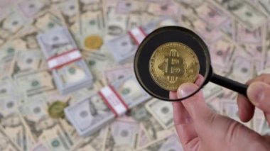 Bitcoin Sikkenin Para Arkaplanındaki Büyüteç ile Macro Görünümü. Elinde bir bitcoin tutan bir kadın, ve onu arıyor ve inceliyor, bir loupe kullanarak. Masaya 100 dolarlık banknotlar serptim..