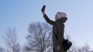 Gülümseyen Kız, Kış Ormanı 'nda bir cep telefonunun sinyalini yakalamaya çalışıyor. Bir bağlantı bulmak için akıllı telefonu açık mavi gökyüzüne doğru uzatan bir kadın. Güneşli açık kış günü. Yapraksız ağaçlar. Doğa.