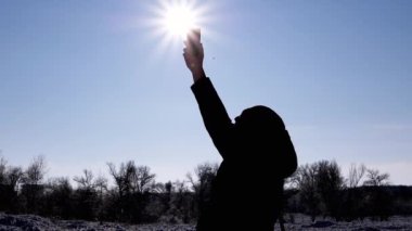 Erkek, Kış Ormanı 'nda bir cep telefonunun sinyalini yakalamaya çalışıyor. Siluetteki adam elini parlak güneşe uzatıyor, mavi gökyüzü sinyal, internet ve bağlantı arıyor. Ufuk. Yaşam Tarzları.