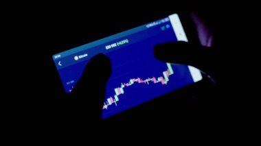 Gece Karanlık Oda 'da Cep Telefonu Uygulamasında Kripto Para Tablosu. Tüccar akıllı telefon ekranına parmağıyla dokunuyor. Dinamik, istatistik, büyüme, düşüş. Borsa. Gelecek fiyatı. Bitcoin.
