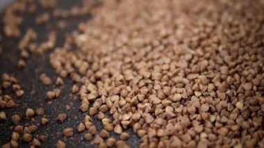 Grains of Buckwheat Fall on a Black Background in Slow Motion. Close up. Raw brown granules and buckwheat particles pour into a pile of buckwheat. Falling buckwheat fills a grainy surface. Texture.