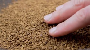 Female Hand Touches with Fingers the Granules of Loose Instant Coffee in Pile. Close up. Hands are holding, sorting through brown particles of coffee beans on a coffee background. Texture, light.