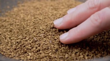 Female Hand Touches with Fingers the Granules of Loose Instant Coffee in Pile. Close up. Hands are holding, sorting through brown particles of coffee beans on a coffee background. Texture, light.