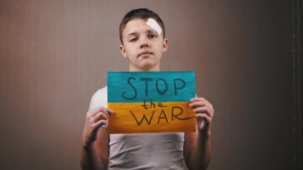 少年碑文とウクライナの旗のポスターを保持する戦争を停止します 彼の目に涙を浮かべて10代の抗議者は ウクライナでの戦争を終わらせるために呼び出します 室内だ 灰色の背景 青い黄色の旗 — ストック動画