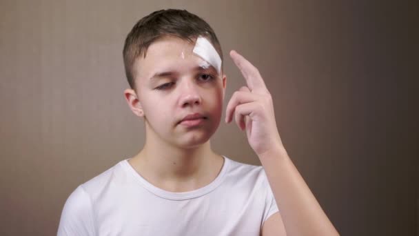 在孤立的灰色背景上 一个头戴粘性石膏的青少年的画像 悲哀的男孩用手触摸额头上的绷带 头部受伤 弹片伤医疗救助 乌克兰战争 — 图库视频影像