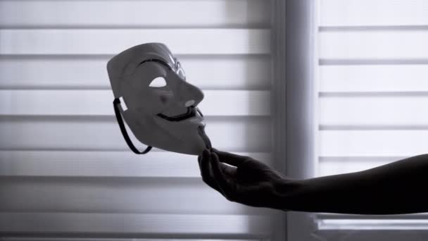 暗い部屋の窓の背景に匿名のマスクを手で保持します 人のアイデンティティを隠すマスクを持つ認識できない人の拡張された手のシルエット スペースのコピー — ストック動画