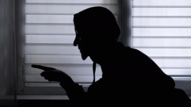 Pencerenin yanında Maskeli Adam 'ın Silueti Yumruğunu Gösteriyor. Tehdit edici, agresif bir hareket. Titreyen yumruk. Yan görüş. Kapüşonlu bir adamın profili. Karanlık oda. Gölge oyunu. İzole edilmiş.