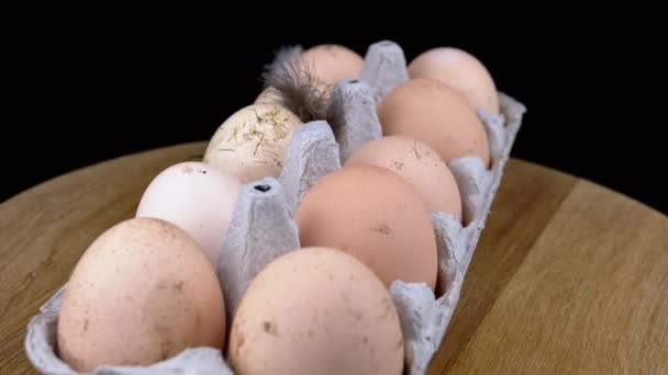 回転する木製の机の上のトレイの茶色の汚い鶏の卵 黒の背景 向きを変えて 羽のついた10個の生の乱雑な卵 エコ製品 タンパク質 段ボール箱 — ストック動画