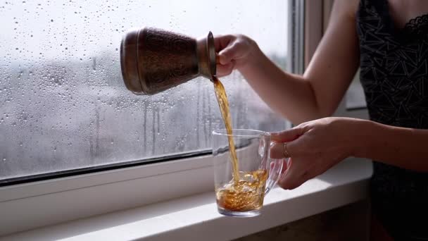 女の子は窓のそばに立っている間にトルココーヒーをカップに準備しました 熱い飲み物 お茶は晴れた雨の日を背景に透明なガラスに注ぎ込まれます 陽射し 朝コーヒーを飲みなさい — ストック動画