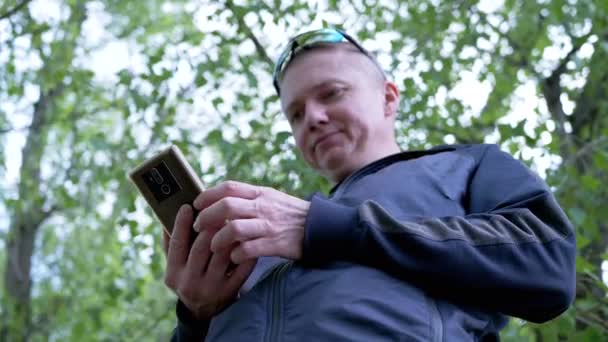 閉じる 幸せな男性自然の森の中でスマートフォンでメッセージを書く 一番下だ 笑顔の観光客 サングラスのサイクリストは オンラインで通信します コンテンツを読みます 屋外レクリエーションを楽しんで — ストック動画