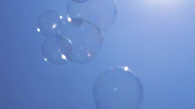 Mavi Gök 'te bir sürü şeffaf parlayan sabun köpüğü uçuşuyor. Temiz mavi arkaplan. Güneş parlıyor, yansıyor. Renkli kabarcıklar hızlı hareket ediyor. Alt manzara. Seçici odaklanma. Doğa arkaplanı.