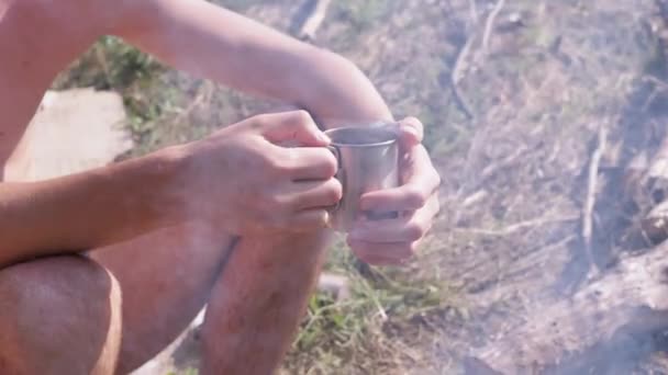 男子在森林里吸烟篝火的背景下喝热咖啡杯 靠近点手 金属杯子 靠近篝火的森林里舒适的气氛 放松点日落了阳光灿烂的夏日 — 图库视频影像