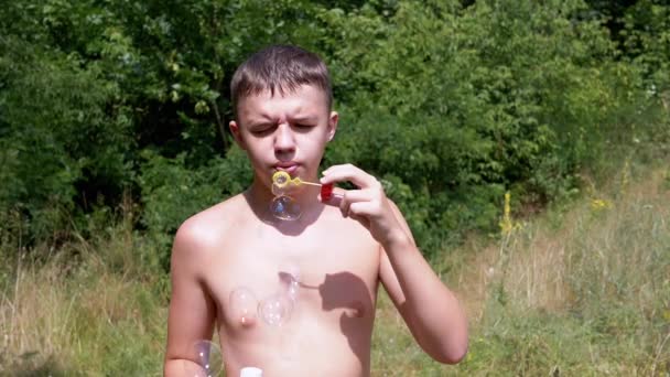 在阳光照射下 一个赤身裸体的男孩在大自然中吹肥皂泡 靠近点一个少年正在森林里放松 吹着五彩斑斓的肥皂泡 模糊的绿色背景 — 图库视频影像
