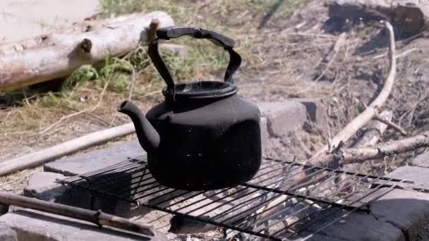 男性は自然の中でホットコーヒーを作るために森で火をつけます 古い沸騰したケトルは 煙の中の金属の格子に浸した 灰と火花を飛んでいる 屋外で バケーション キャンプ — ストック動画