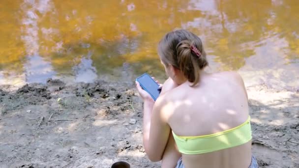 自然の川の近くに座っているスマートフォンにメッセージを書く少女 バックビュー 濡れた砂の上に座っている黄色い水着の少女は 水の背景に対してSmsを書きます 休憩する アウトドア — ストック動画