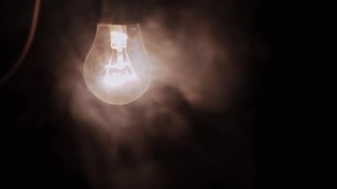 Siyah Arkaplanda Kalın Duman Altında Kabloyla Parlayan Bir Ampul. Tozlu, aktif, eski Edison tungsten ampulü dumanlı karanlık bir odada sarı bir ışık parlıyor. Görüntü. Doku.