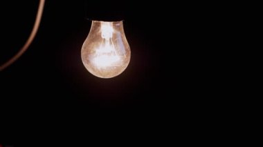 Siyah Arkaplanda Kablosu Olan Bir Asılı, Parlayan Ampul. Tozlu, eski bir Edison tungsten ampulü karanlık odada loş sarı bir ışık parlıyor. Titreşiyor. Elektrik tasarrufu. Yavaş çekim.