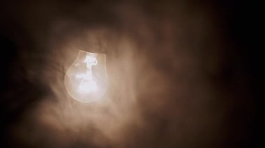 Siyah Arkaplanda Kalın Duman Altında Bir Asılı, Parlayan Ampul. Sallanan, Tozlu, Edison tungsten ampulünü açmış dumanlı karanlık bir odada sarı bir ışık parlıyor. Bulanık hareket. Doku. Sis.
