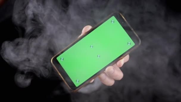 在黑烟中手握带有绿色屏幕的手机 手动旋转一个带有空白屏幕的智能手机 彩色键 在浓密的烟雾中模拟在空旷的空间 — 图库视频影像