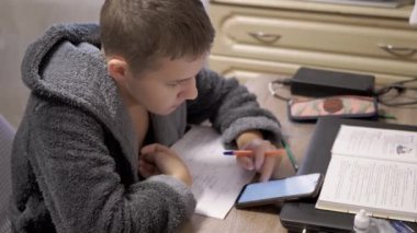 Masada oturan, akıllı telefon kullanarak ödev yapan, bornozlu ciddi bir genç. Öğrenci çocuk dikkatlice bir web sayfası okuyor, ekranda parmağıyla kaydırıyor. Çevrimiçi öğrenim, evde eğitim.