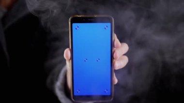Smoke 'ta Siyah Arkaplanda Mavi Ekranlı Cep Telefonu Tutmak. El, bir akıllı telefonu boş bir ekranla döndürür, kroma anahtarı, kalın bir duman bulutu içinde boş uzayda taklit eder. Doku.