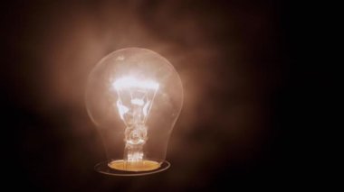 Siyah Arkaplanda Kalın Duman 'da Parlayan Bir Yanan Ampul. Kapatın. Tozlu, açık Edison tungsten ampulü dumanlı karanlık odada sarı bir ışık parlıyor. Bulanık hareket. Doku. Sis.