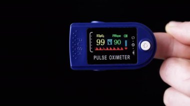 Kadın, Nabız ve Oksijen Doygunluğunu Evde Nabız Oksimetresi kullanarak Ölçüyor. Yakın plan. Siyah arka plan. Cihazın üzerinde sensörler ve göstergeler olan bir arama. Kalp atışlarının, nabzının, kalp atışlarının ölçümü.