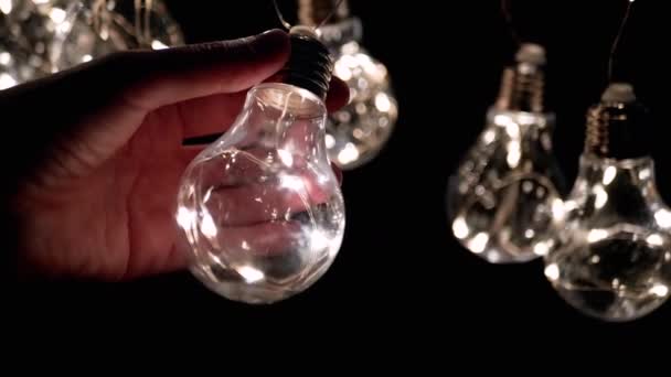 手握一个灯泡在一个发光灯泡的花园的背景上 圣诞装饰 复古风格的老式电灯泡照亮了这个空间 黑暗房间 — 图库视频影像