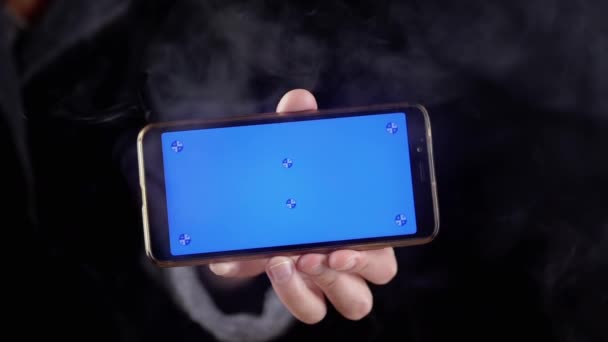 在黑烟中手握带有蓝色屏幕的手机 手动旋转一个带有空白屏幕的智能手机 彩色键 在浓密的烟雾中模拟在空旷的空间 — 图库视频影像