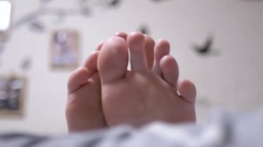 Yaklaş, Yatak odasındaki yatakta pijamayla yorgun bacakları hareket ettiren kişi. Çocuk ayak parmaklarını oynatıyor. Jimnastik. Sıkıştırma, uzuvların ve ayakların çözülmesi. Kramplardan, stresten, yorgunluktan kurtuldum. Rahatlama.