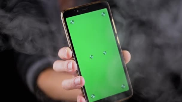在黑烟中手握带有绿色屏幕的手机 手动旋转一个带有空白屏幕的智能手机 彩色键 在浓密的烟雾中模拟在空旷的空间 — 图库视频影像