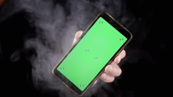 煙の黒い背景に緑色のスクリーンが付いている携帯電話を握る手 ハンドは 空白の画面 クロマキーでスマートフォンを回転させ 煙の厚い雲の中で空のスペースでモックアップします テクスチャー — ストック動画