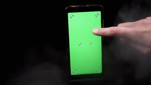 煙の黒い背景に緑色のスクリーンが付いている携帯電話を握る手 指はクロマキー メーカーが付いている空白のタッチスクリーンのスマートフォンに触れます 空っぽの空間 煙の厚い雲だった テクスチャー ブロール — ストック動画