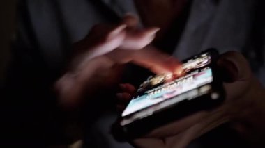 Karanlık bir ortamda akıllı telefon ekranında içerikte süzülen ellerin yakın çekim görüntüsü. Parmaklar ekranın yumuşak parıltısıyla aydınlanan arayüzde zarifçe geziniyor. Uygun.