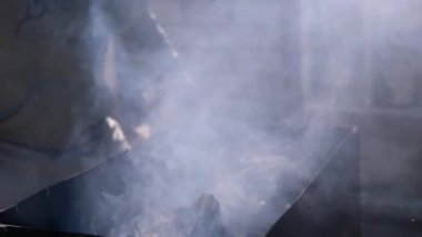 Kürekle yanan kömürleri yelpazeleyen bir adam, közleri hayata döndürerek beyaz bir duman bulutu yaratıyor..