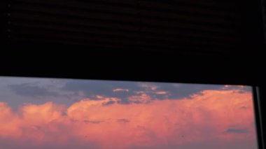 Pembe ve turuncu günbatımı bulutları pencere perdelerindeki bir boşluktan görülür ve huzurlu ve huzurlu bir atmosfer yaratır. Gökyüzünün canlı renkleri büyüleyici bir görüntü sağlar..