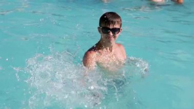 Güneş gözlüğü takan bir genç yazın tadını çıkarıyor, canlı bir turkuaz havuzda neşeyle sıçrıyor, ferahlatıcı su püskürtüleri yaratıyor. Kaygısız ruh hali yaz eğlencesinin özünü yakalar..