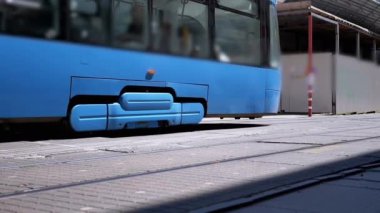 İstasyona mavi bir tramvay yaklaşıyor ve uygun bir toplu taşıma aracı sunuyor. Bu sahne kentsel bağlantıyı resmediyor.