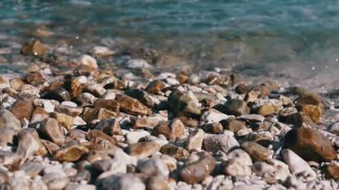 Plajda ıslak çakıl taşlarının yakın çekimlerinde, yumuşak dalgalar üzerlerini kaplıyor. Temiz su gün ışığında parlıyor..