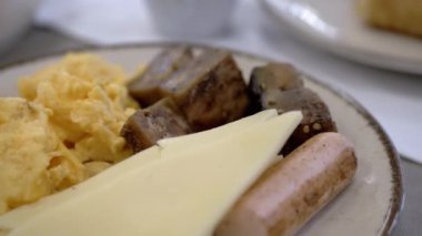 Çırpılmış yumurta, peynir, sosis ve ızgara sebzeli kahvaltı tabağının detaylı görüntüsü. Yakın çekim yemeğin dokusunu ve rengini ön plana çıkarıyor, bu da onu mükemmel bir görsel hale getiriyor.