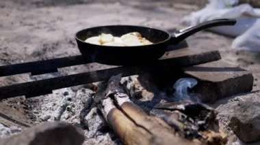 Kamp ateşinde tavada kızartılmış patates. Yemek pişerken buhar yükselir ve kırsal bir açık hava yemek sahnesi yaratır. Arka planda beyaz bir çanta duruyor..