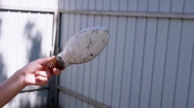 Bir el, yanmış, kirli bir ampulü ahşap bir çite karşı tutar. Bu görüntü atık kavramlarını, eskimeyi ve tehlikeli maddelerin düzgün bir şekilde imha edilmesine duyulan ihtiyacı temsil ediyor..