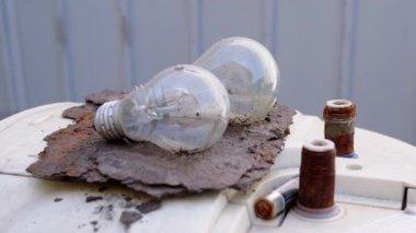 İki yanmış ampul paslı bir metal parçasının üzerinde duruyor, atığı ve eskiyi sembolize ediyor. Arka plandaki beyaz metal duvar sahneye katıksız bir karşıtlık ekler.